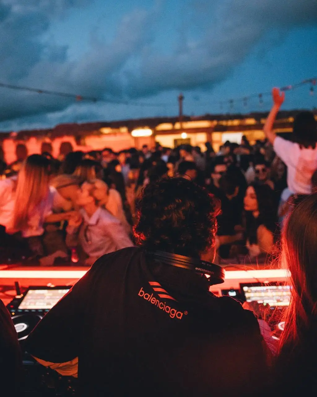 A DJ at the decks at dusk, headphones round the neck, a lit crowd filling the terrace behind